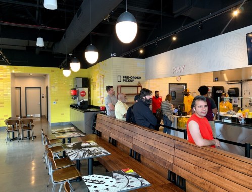 A bright, modern restaurant interior with customers at the counter and staff in yellow shirts. Wood seating in foreground.