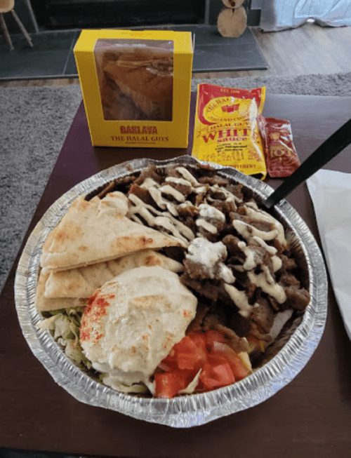A foil container filled with meat, rice, salad, pita, and sauces, alongside baklava and condiments.
