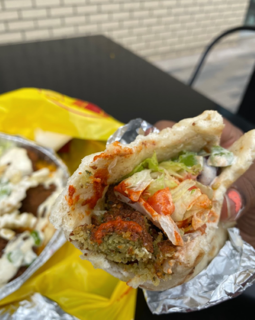 A hand holding a stuffed pita with falafel, lettuce, and sauce, with a yellow takeout bag in the background.