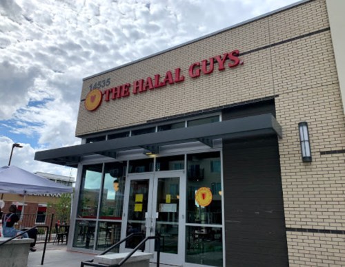 Exterior of The Halal Guys restaurant, featuring a modern design and large windows under a cloudy sky.