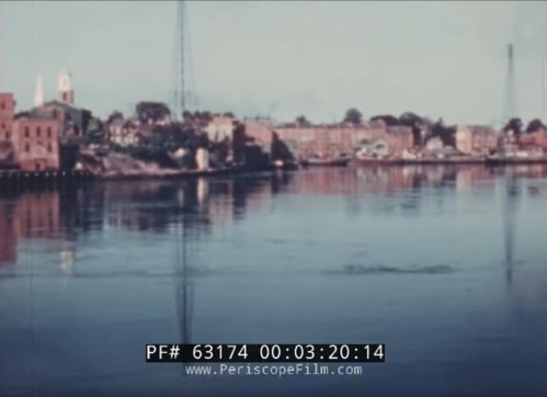 A serene river scene with buildings and a tower reflected in the water, under a clear sky.