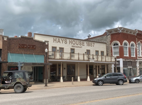 Historic Hays House restaurant with a balcony, flanked by brick buildings under a cloudy sky.