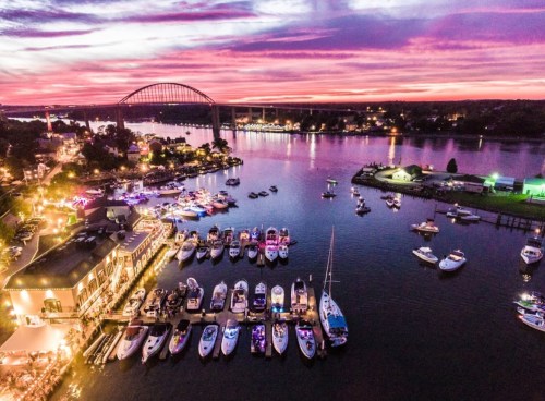 Aerial view of a vibrant waterfront at sunset, with boats docked and colorful lights reflecting on the water.