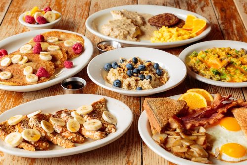 A spread of breakfast dishes including pancakes, oatmeal, eggs, bacon, and fresh fruit on wooden table.