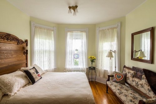 Cozy bedroom with a bed, vintage decor, large windows, and a small seating area with a mirror and flowers.