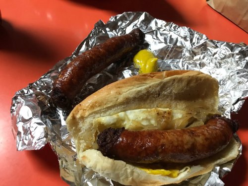 A sausage sandwich with grilled onions and mustard, alongside an extra sausage on a foil-lined plate.