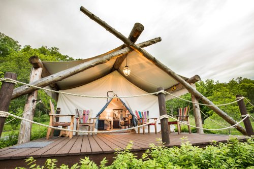 A cozy glamping tent with wooden supports, surrounded by greenery and featuring colorful chairs on a wooden deck.