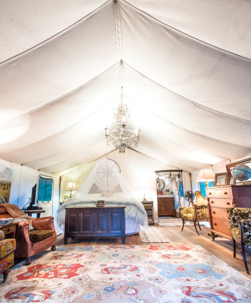 A spacious tent interior featuring a bed, chandelier, vintage furniture, and a colorful rug.
