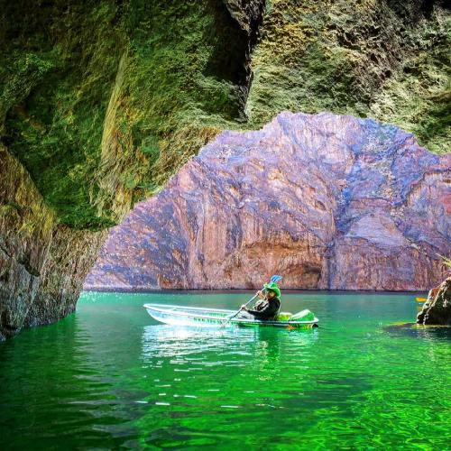 A person in a kayak paddles through a serene, green lake surrounded by towering rock formations and cliffs.