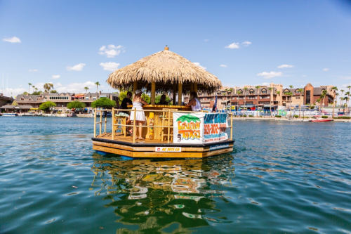 A thatched-roof floating hut on water, with people enjoying the view and a colorful banner on the side.
