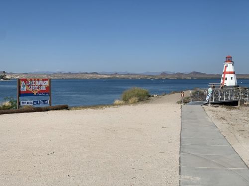 A sandy beach with a lighthouse and a sign for Lake Havasu State Park, overlooking a calm lake and distant mountains.