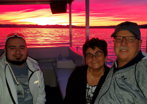 Three people smiling on a boat at sunset, with vibrant pink and orange skies reflecting on the water.