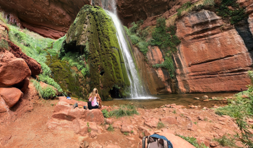 A person sits by a moss-covered waterfall in a red rock canyon, surrounded by lush greenery and a serene pool.