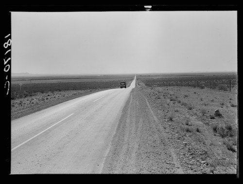 A long, empty road stretches through a barren landscape, with a single vehicle in the distance under a cloudy sky.