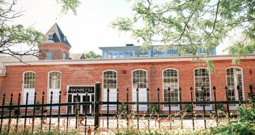A brick building with large windows and a tower, featuring a sign that reads "SAVAGE HILL," surrounded by greenery.