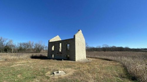 Ruins of a stone building stand in a grassy field under a clear blue sky, surrounded by sparse trees.