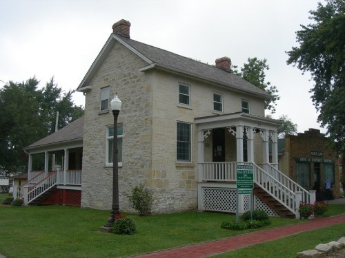 Historic stone house with a porch, surrounded by green grass and flowers, under a cloudy sky.
