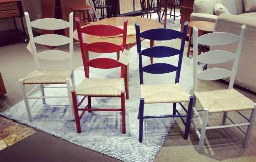 Four colorful chairs in white, red, and blue with woven seats, arranged on a patterned rug in a furniture store.