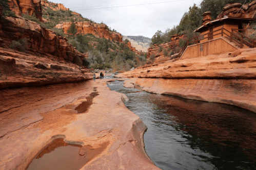 A serene river flows through red rock formations, with people walking along the banks in a scenic canyon landscape.