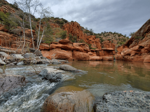 A serene river scene with rocky banks, lush greenery, and red rock formations under a cloudy sky.