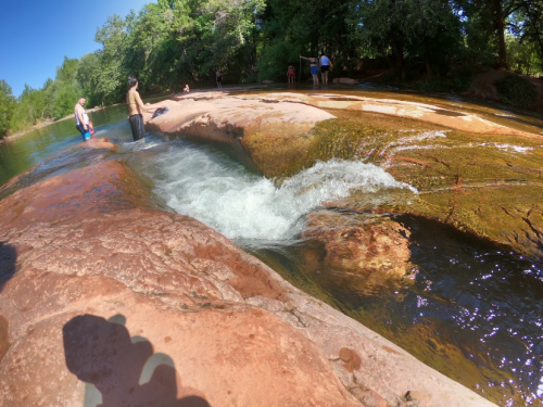 People enjoying a sunny day by a flowing river with smooth rocks and lush greenery in the background.