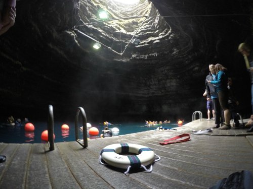 A cavern with a circular opening above, featuring people swimming in blue water and a lifebuoy on a wooden dock.