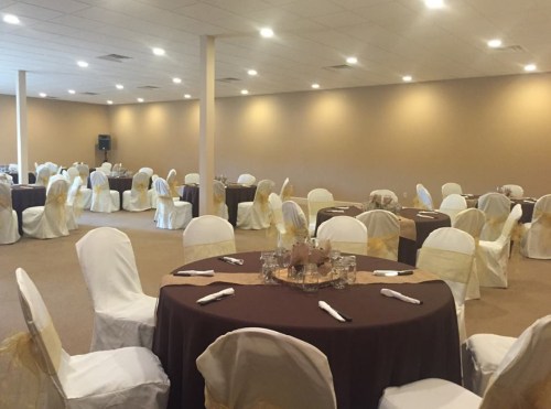 A spacious event room with round tables dressed in brown and white, surrounded by chairs with gold sashes.