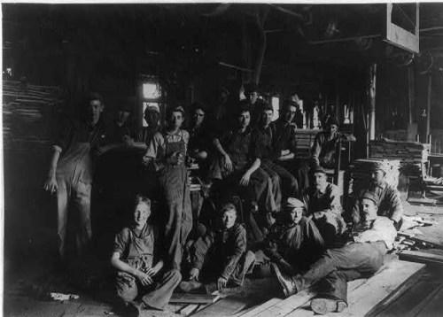 A group of workers, including men and boys, poses inside a dimly lit workshop surrounded by wooden materials.