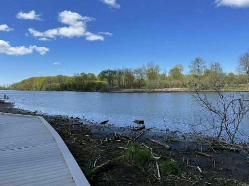 A serene river scene with a wooden walkway, surrounded by trees and a clear blue sky with scattered clouds.