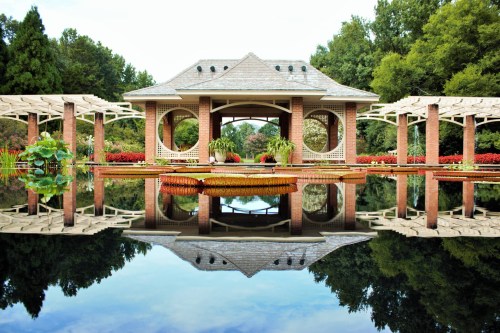 A serene garden pavilion reflected in calm water, surrounded by lush greenery and colorful flowers.