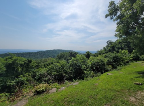 A scenic view of lush green hills under a blue sky, with distant mountains and a hint of water in the background.