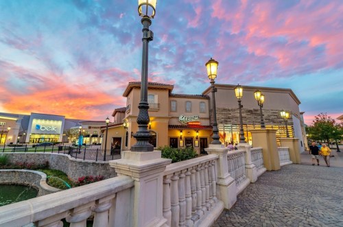 A picturesque shopping area at sunset, featuring a restaurant and retail stores under a colorful sky.