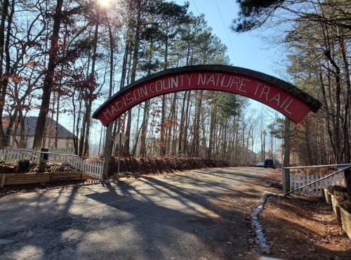 Archway sign for Madison County Nature Trail, surrounded by trees and a clear blue sky. Road leads into the trail.