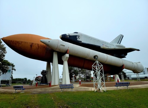 A space shuttle mounted on a large rocket display, set in a park with benches and trees in the background.