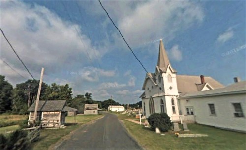 A quiet rural road lined with trees, featuring a white church with a tall steeple and a few nearby buildings.