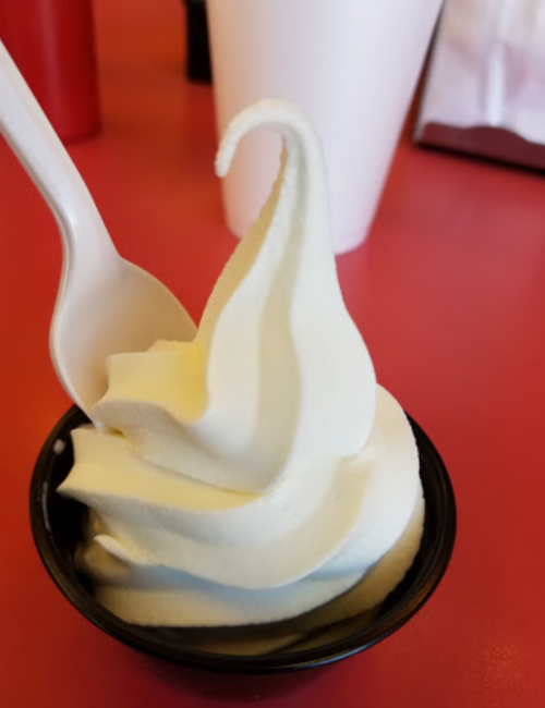 A cup of soft-serve ice cream with a swirl, accompanied by a plastic spoon, on a red table.