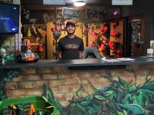 A smiling employee stands behind a counter in a themed rental shop, surrounded by colorful toy guns and jungle decor.