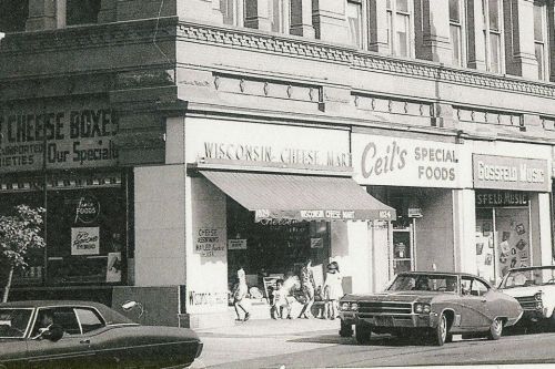 Black and white photo of a street scene featuring shops, including "Wisconsin Cheese Mart" and "Ceil's Special Foods."