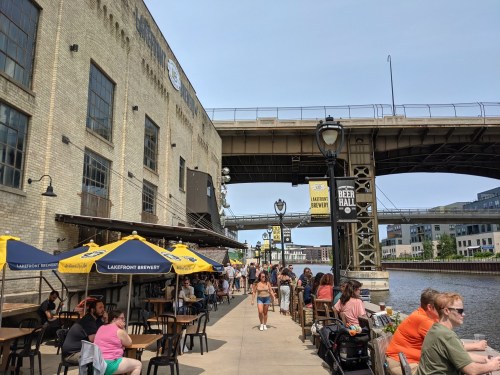 A bustling riverside scene with people dining under umbrellas, next to a brewery and a bridge overhead.