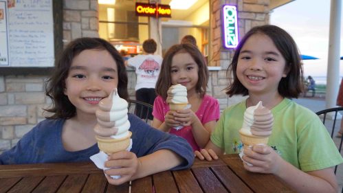 Three girls smile while holding ice cream cones at an outdoor stand, with a neon "Open" sign in the background.