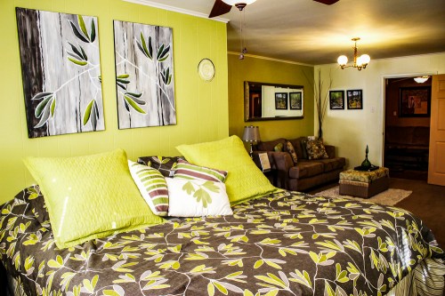 Cozy bedroom with green walls, floral bedding, and artwork, featuring a sofa and decorative plants in a warm atmosphere.