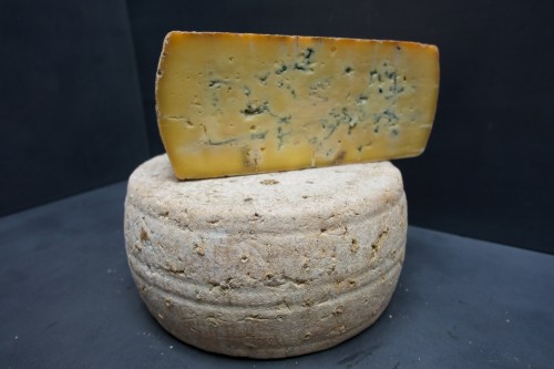 A round wheel of cheese topped with a wedge of blue cheese against a dark background.