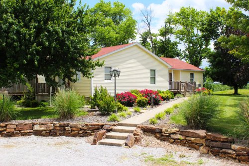 A charming yellow house with a red roof, surrounded by greenery and colorful flowers, set against a blue sky.