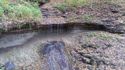 A small waterfall cascades over rocky terrain, surrounded by autumn leaves and greenery.