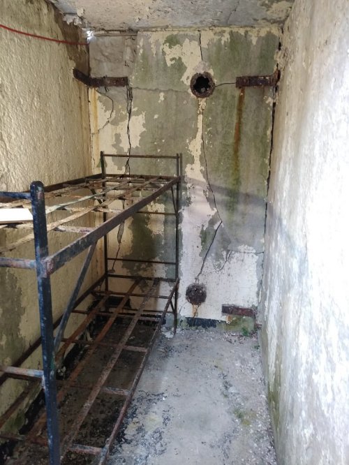A dimly lit, empty room with cracked walls, a rusty metal bed frame, and visible signs of dampness.