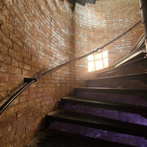 A spiral staircase with wooden steps and a brick wall, illuminated by sunlight through a small window.