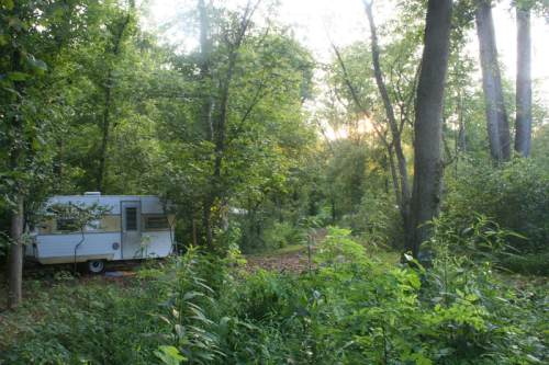A white camper nestled among lush green trees and foliage, with a dirt path leading into the woods at sunset.
