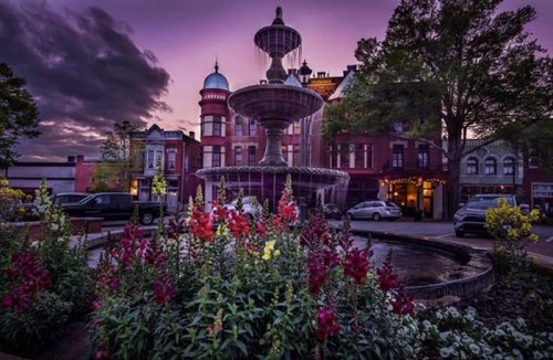 A fountain surrounded by colorful flowers at dusk, with historic buildings and a cloudy sky in the background.