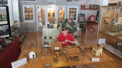 Two men in a museum, one seated at a table with miniature buildings, the other standing behind him.