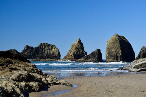Rocky formations rise from the ocean under a clear blue sky, with gentle waves lapping at the sandy beach.